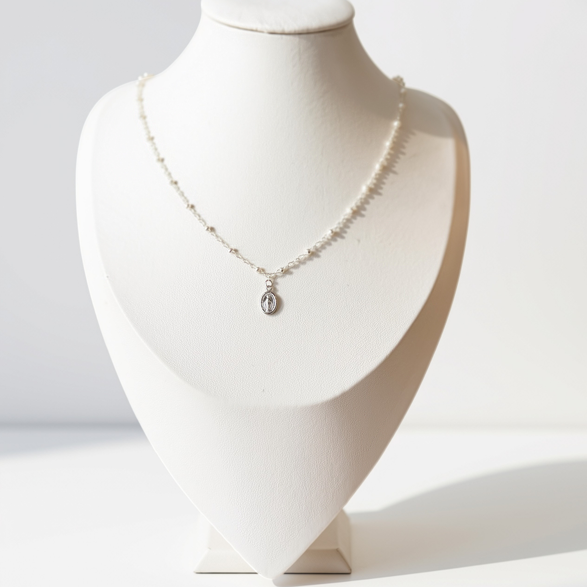 A delicate silver necklace with a small oval pendant displayed on a white jewelry bust against a bright white background.