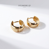 A pair of gold huggie hoop earrings with a textured, basketweave-like pattern on the front and polished edges, displayed on a clean white surface with soft shadows. The brand name 'SYCAMORE LEE JEWELRY' is printed at the top.