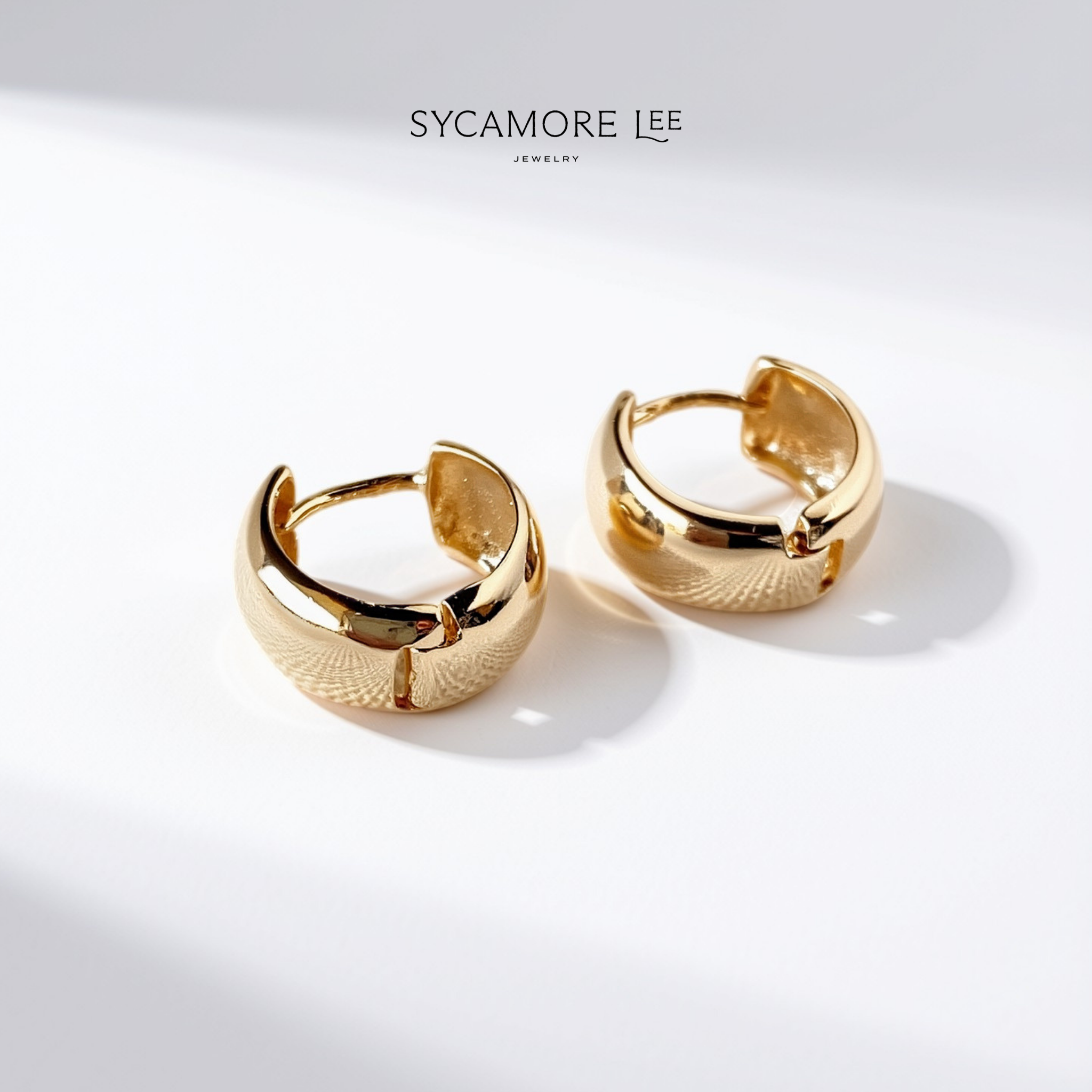 A pair of gold huggie hoop earrings with a textured, basketweave-like pattern on the front and polished edges, displayed on a clean white surface with soft shadows. The brand name 'SYCAMORE LEE JEWELRY' is printed at the top.