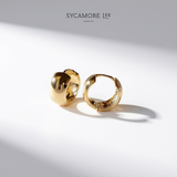 A pair of gold huggie hoop earrings with textured and polished finishes, displayed on a clean white surface with dramatic diagonal shadows. The brand name 'SYCAMORE LEE JEWELRY' is printed at the top center.