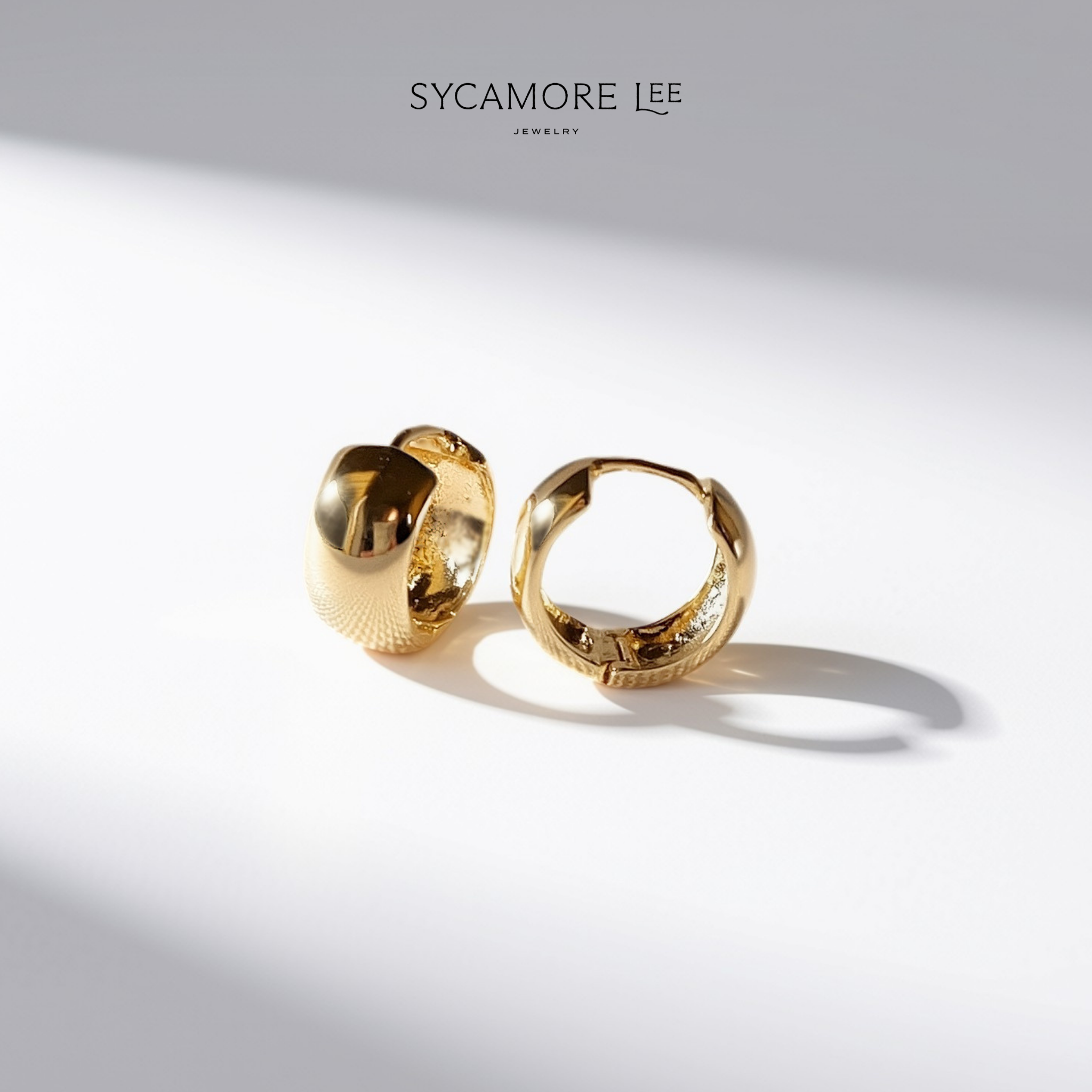 A pair of gold huggie hoop earrings with textured and polished finishes, displayed on a clean white surface with dramatic diagonal shadows. The brand name 'SYCAMORE LEE JEWELRY' is printed at the top center.