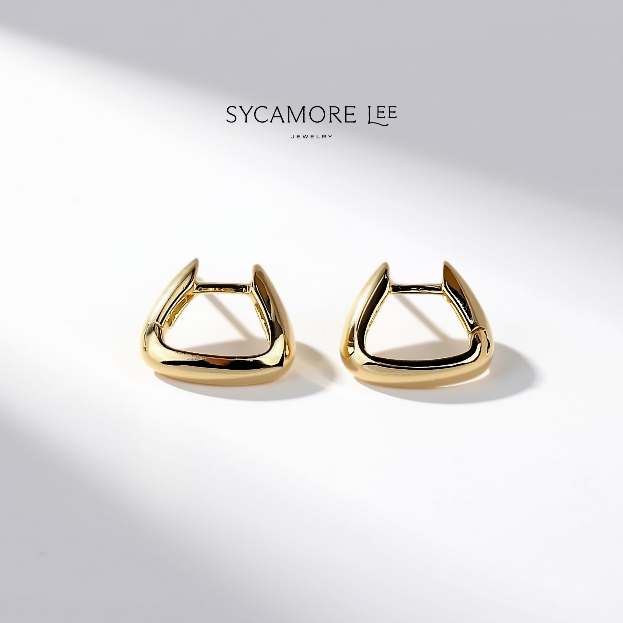 A pair of gold-toned geometric hoop earrings with a triangular open-frame design, displayed on a clean white surface with soft shadows. The brand name 'SYCAMORE LEE JEWELRY' is printed above the earrings.