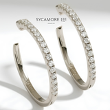 A pair of silver-toned hoop earrings with a row of clear pavé-set round stones, shown on a white background with the brand name 'SYCAMORE LEE JEWELRY' printed above.