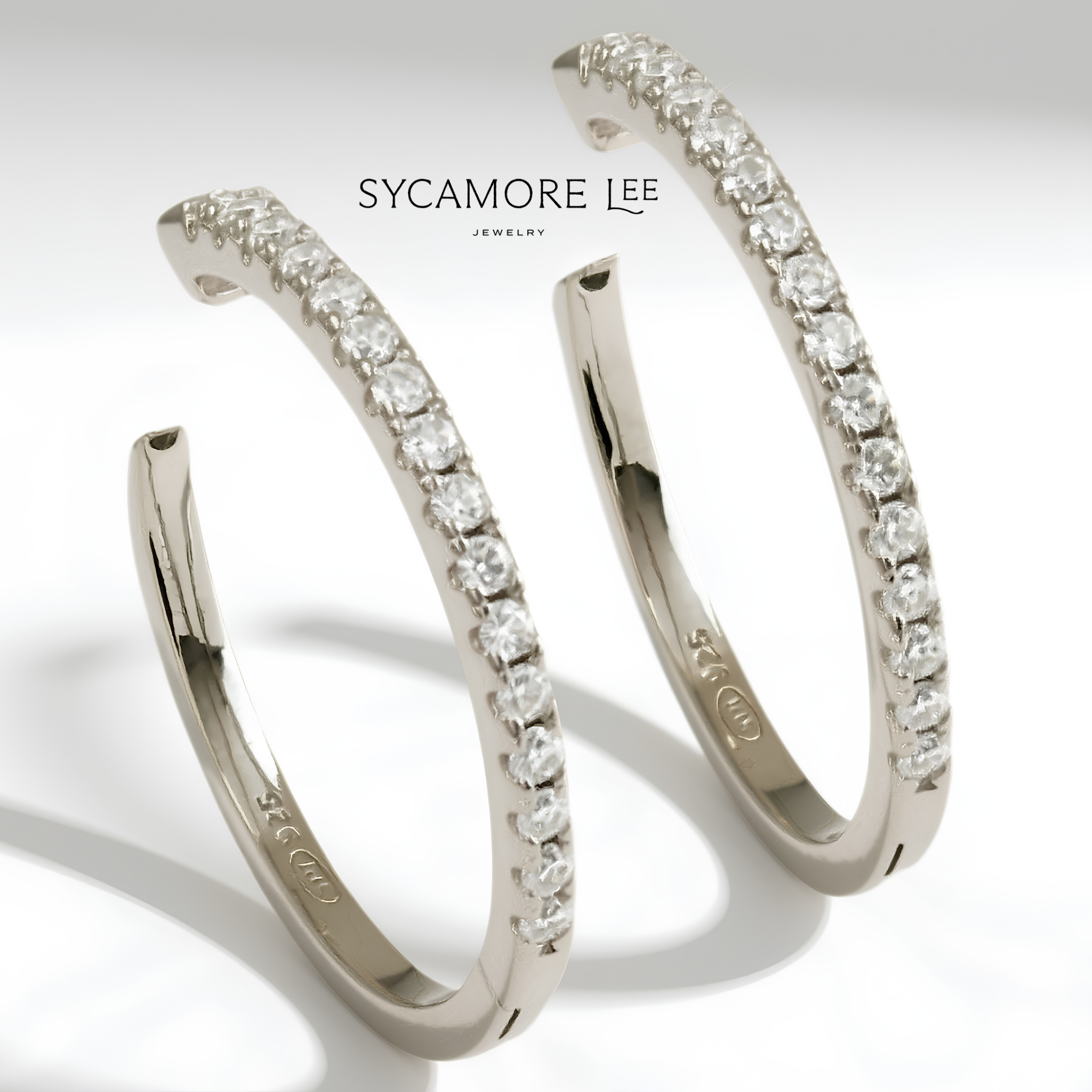 A pair of silver-toned hoop earrings with a row of clear pavé-set round stones, shown on a white background with the brand name 'SYCAMORE LEE JEWELRY' printed above.