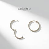 A pair of silver hoop earrings with textured beaded edges, displayed on a clean white background with soft diagonal shadows. The brand name 'SYCAMORE LEE' is printed in black at the top center.
