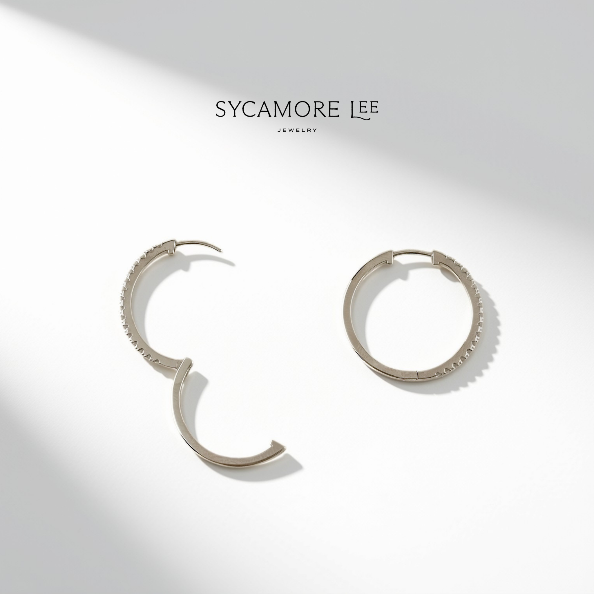 A pair of silver hoop earrings with textured beaded edges, displayed on a clean white background with soft diagonal shadows. The brand name 'SYCAMORE LEE' is printed in black at the top center.