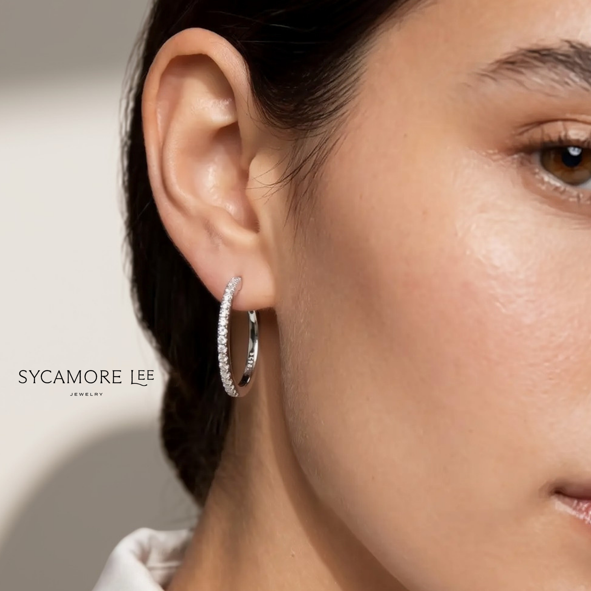 A close-up of a model's ear wearing a thin silver hoop earring encrusted with small clear stones. The brand name 'SYCAMORE LEE' is printed on the left side of the image against a clean, light beige background.