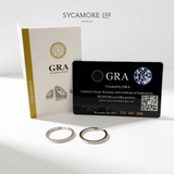 Two silver hoop earrings displayed in front of branded packaging on a clean white background. The set includes a white and gold Sycamore Lee Jewelry insert card, a black GRA warranty card, and two silver hoops, one with a small row of clear stones.