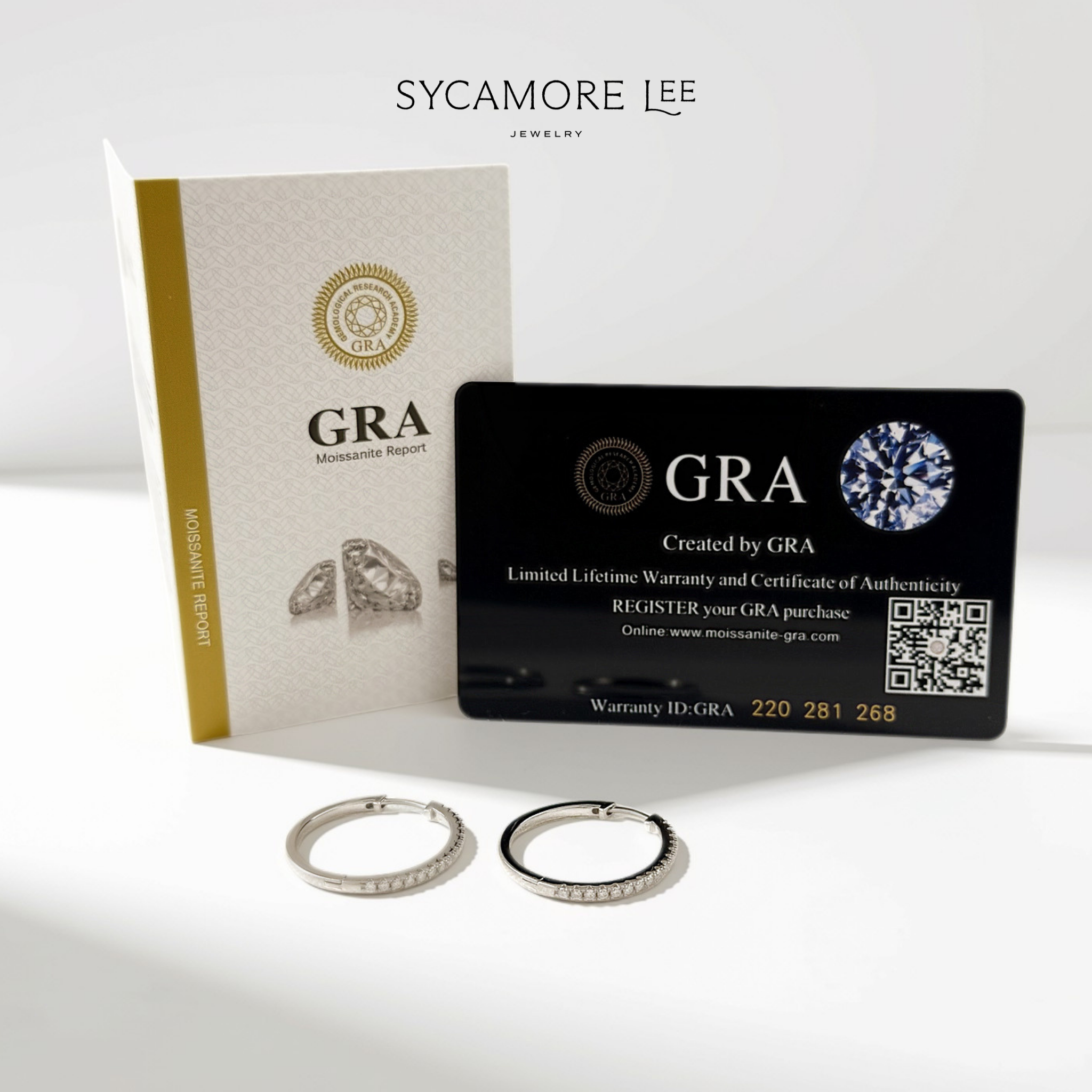 Two silver hoop earrings displayed in front of branded packaging on a clean white background. The set includes a white and gold Sycamore Lee Jewelry insert card, a black GRA warranty card, and two silver hoops, one with a small row of clear stones.