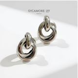 A pair of polished silver-toned hoop earrings with a twisted knot design, displayed on a clean white surface with soft shadows. The brand name 'SYCAMORE LEE JEWELRY' is printed at the top.