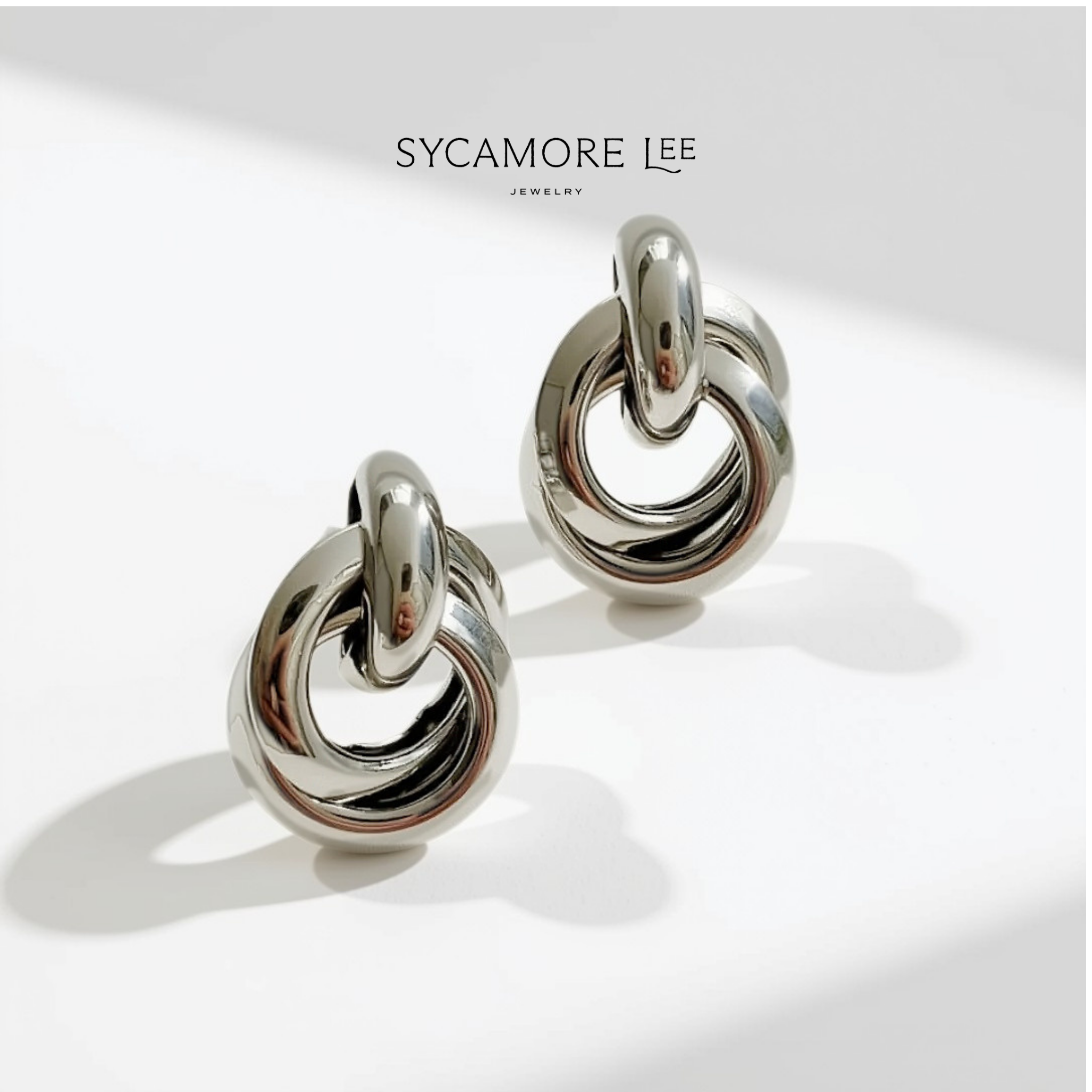 A pair of polished silver-toned hoop earrings with a twisted knot design, displayed on a clean white surface with soft shadows. The brand name 'SYCAMORE LEE JEWELRY' is printed at the top.