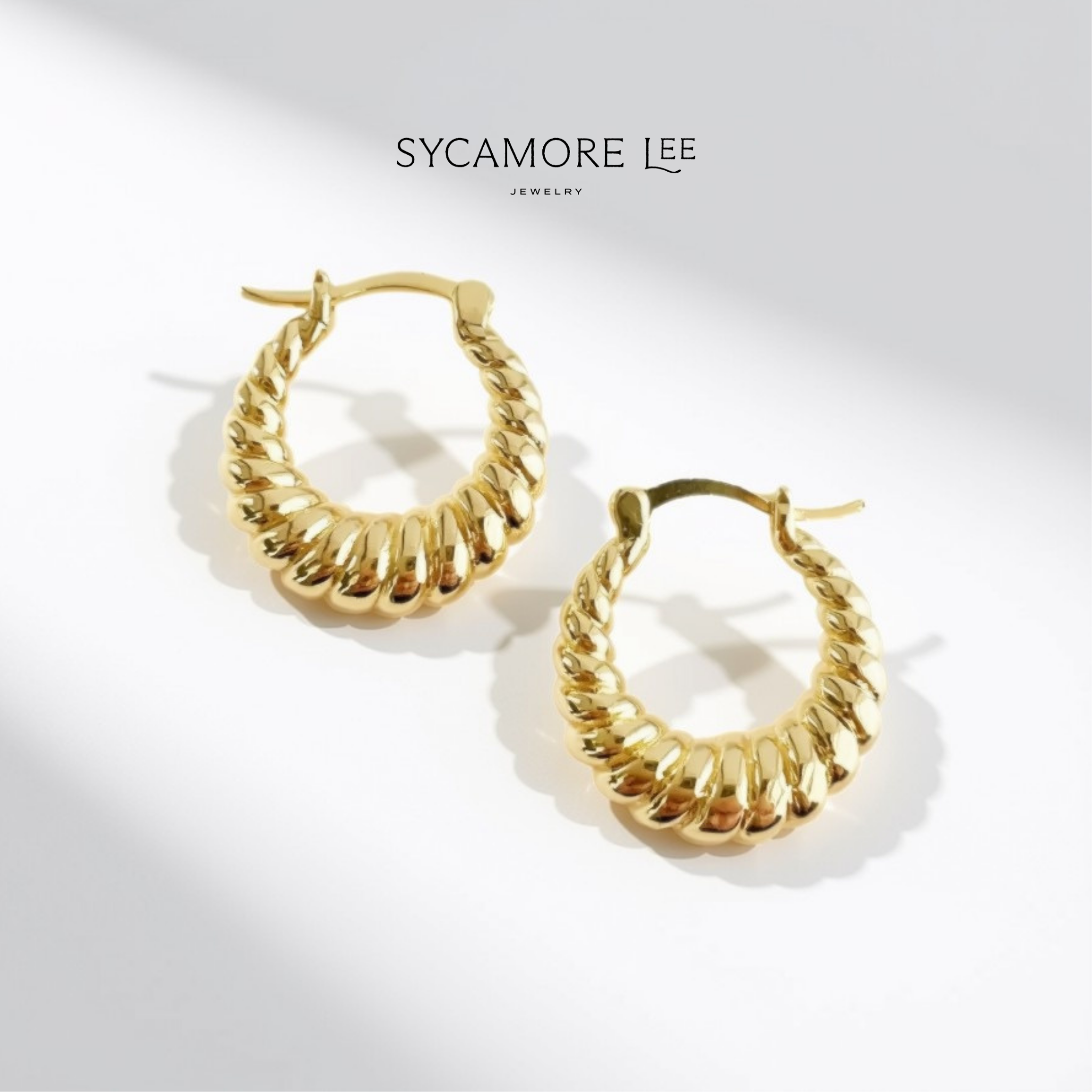 A pair of gold hoop earrings with a textured, ridged outer design and a twisted rope-like inner band, displayed on a clean white background with soft shadows. The brand name 'SYCAMORE LEE JEWELRY' is printed at the top.