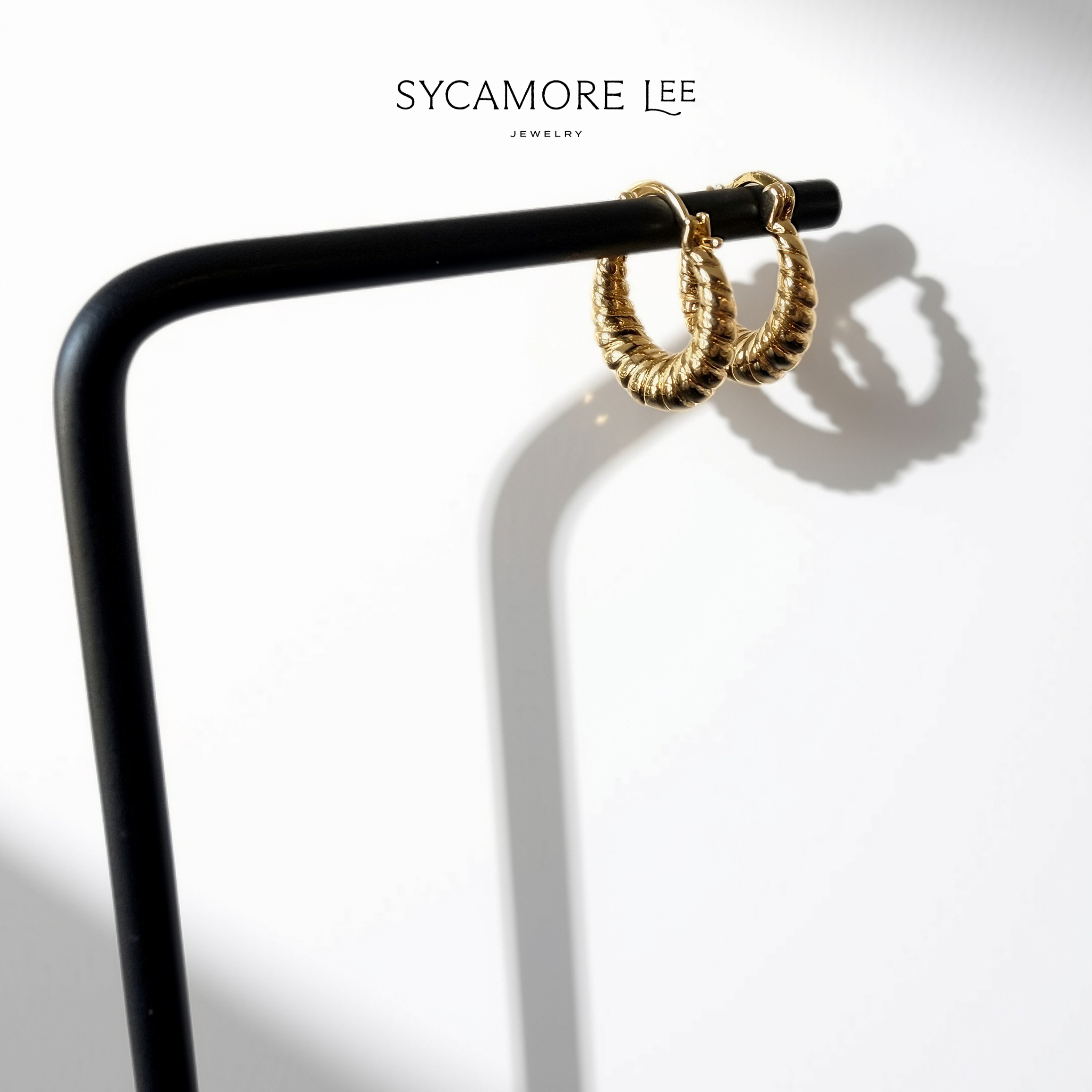 Two textured gold hoop earrings displayed on a black metal stand against a white background, with the brand name 'SYCAMORE LEE JEWELRY' printed at the top.