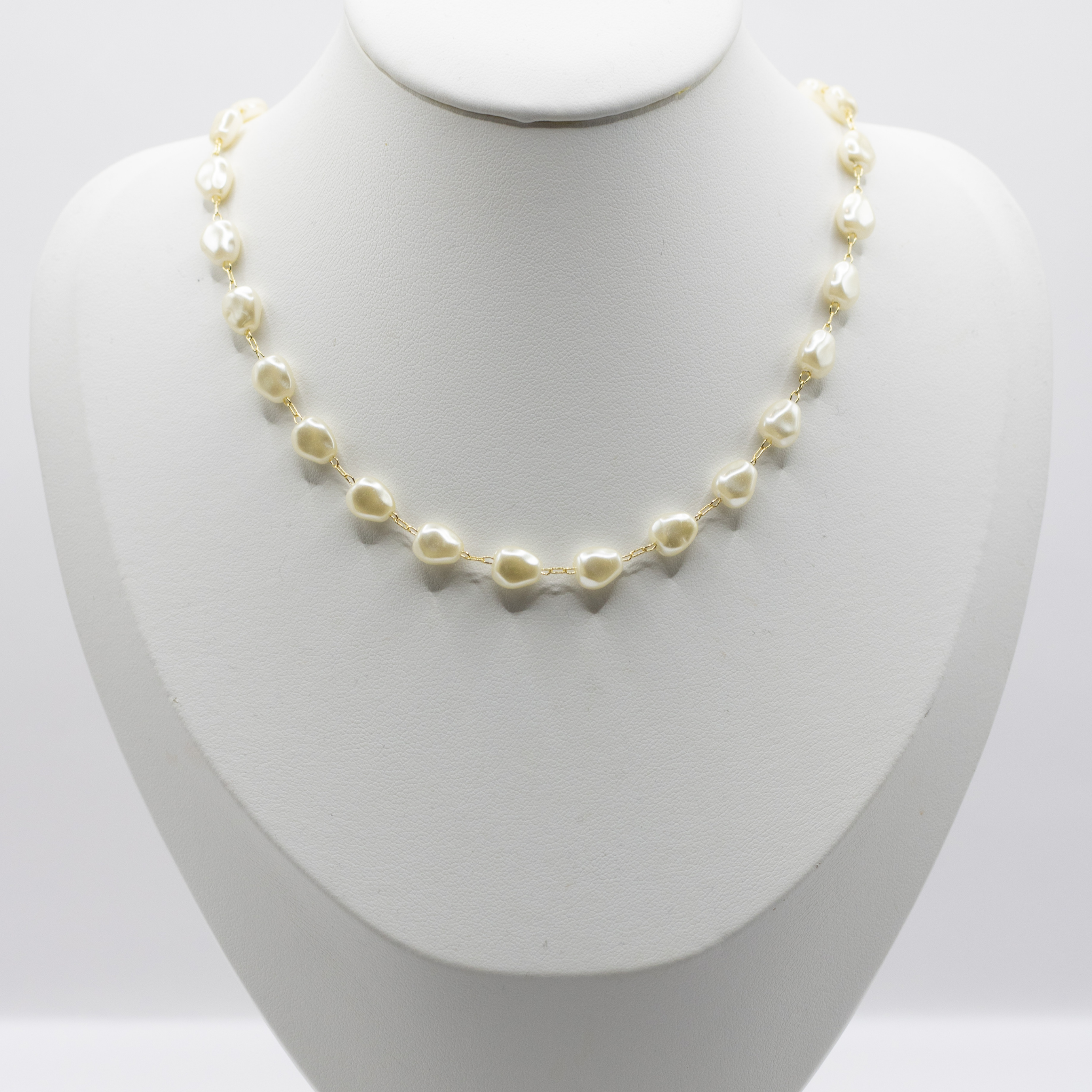 A delicate necklace made of small irregular cream-colored pearls on a gold chain, displayed on a white jewelry bust against a light gray background.