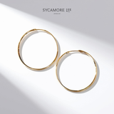 A pair of gold hoop earrings displayed on a clean white background with artistic diagonal shadows. The brand name 'SYCAMORE LEE JEWELRY' is printed at the top center.