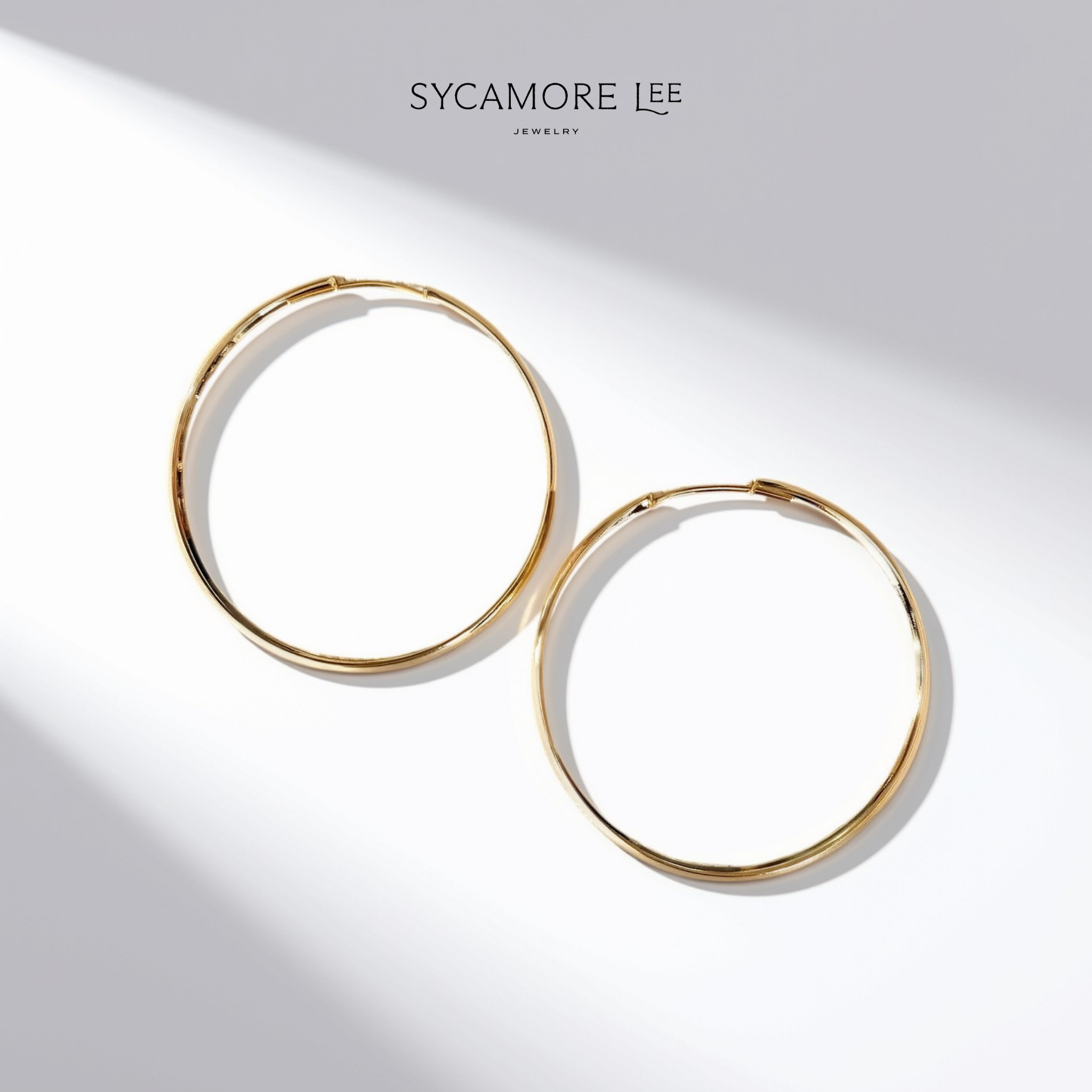 A pair of gold hoop earrings displayed on a clean white background with artistic diagonal shadows. The brand name 'SYCAMORE LEE JEWELRY' is printed at the top center.
