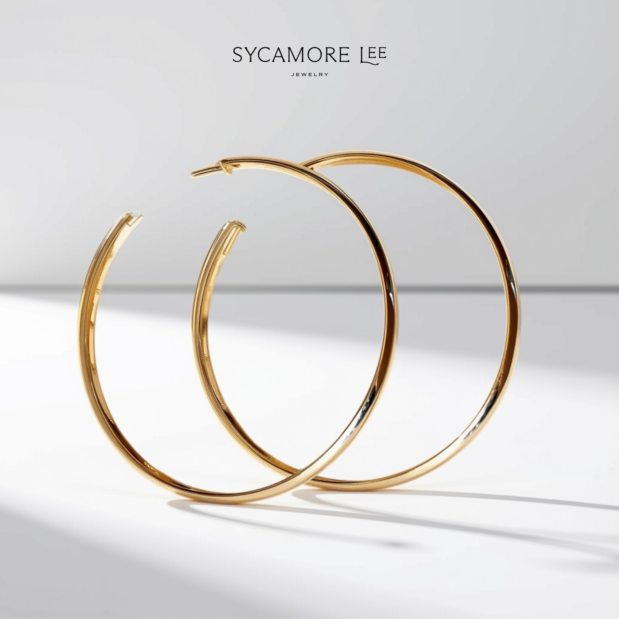 A pair of gold hoop earrings with a polished finish, displayed on a clean white background with a soft shadow. The brand name 'SYCAMORE LEE' is printed at the top center.