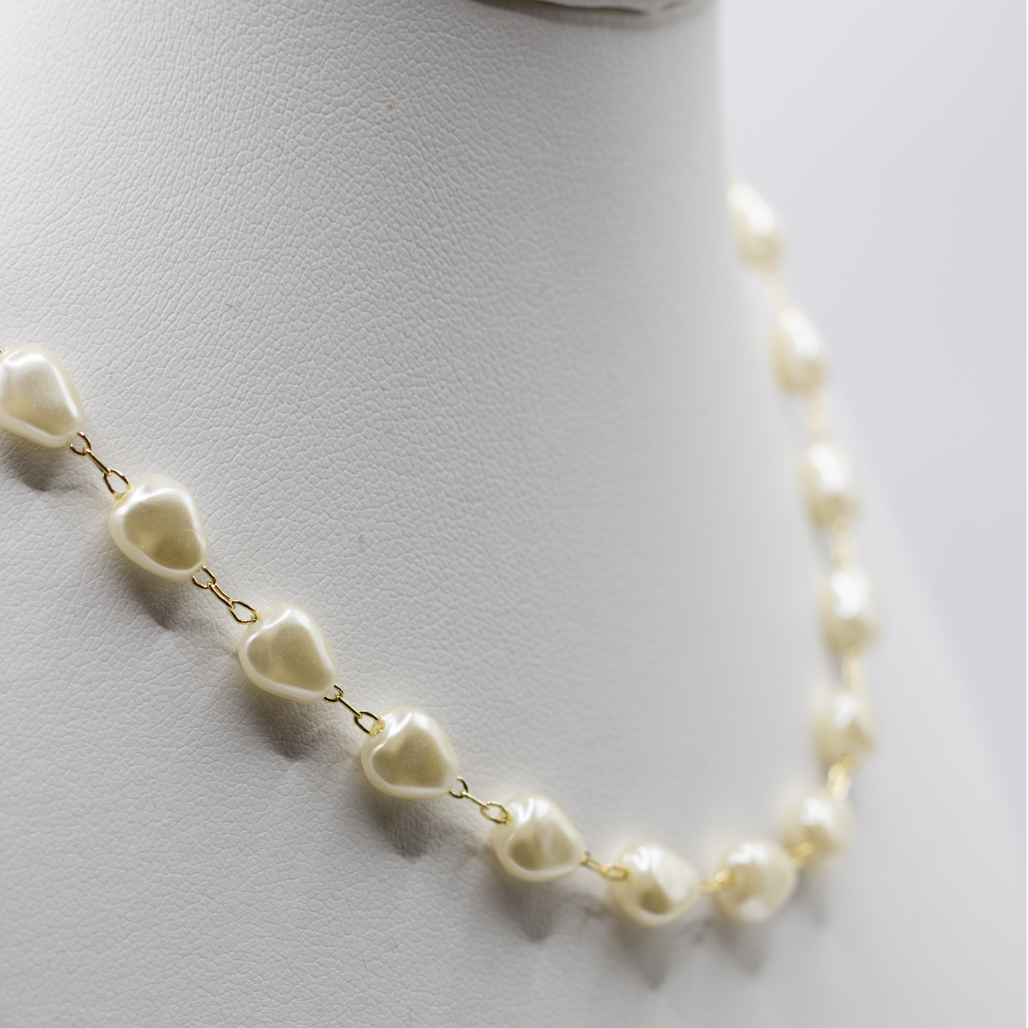 A close-up of a necklace with irregular cream-colored pearls connected by small gold chains, displayed on a white mannequin bust.