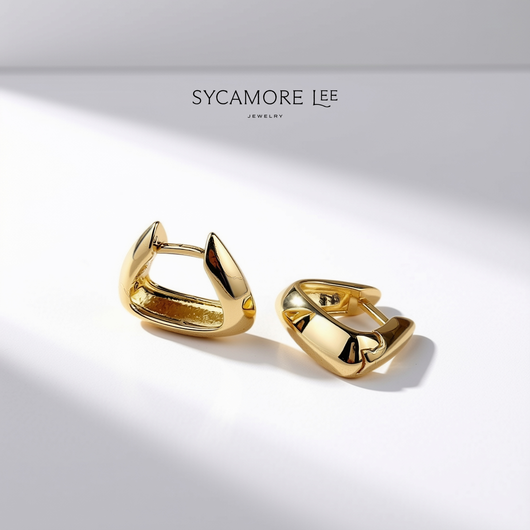 A pair of gold huggie hoop earrings with a polished, sculptural organic shape, displayed on a clean white surface with artistic diagonal shadows. The brand name 'SYCAMORE LEE JEWELRY' is printed at the top center.