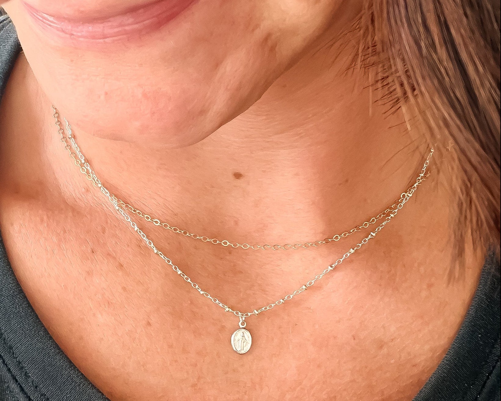 A close-up lifestyle shot of a woman wearing a delicate double-layered silver necklace with a small miraculous medal pendant on a sterling silver delicate cable chain. 