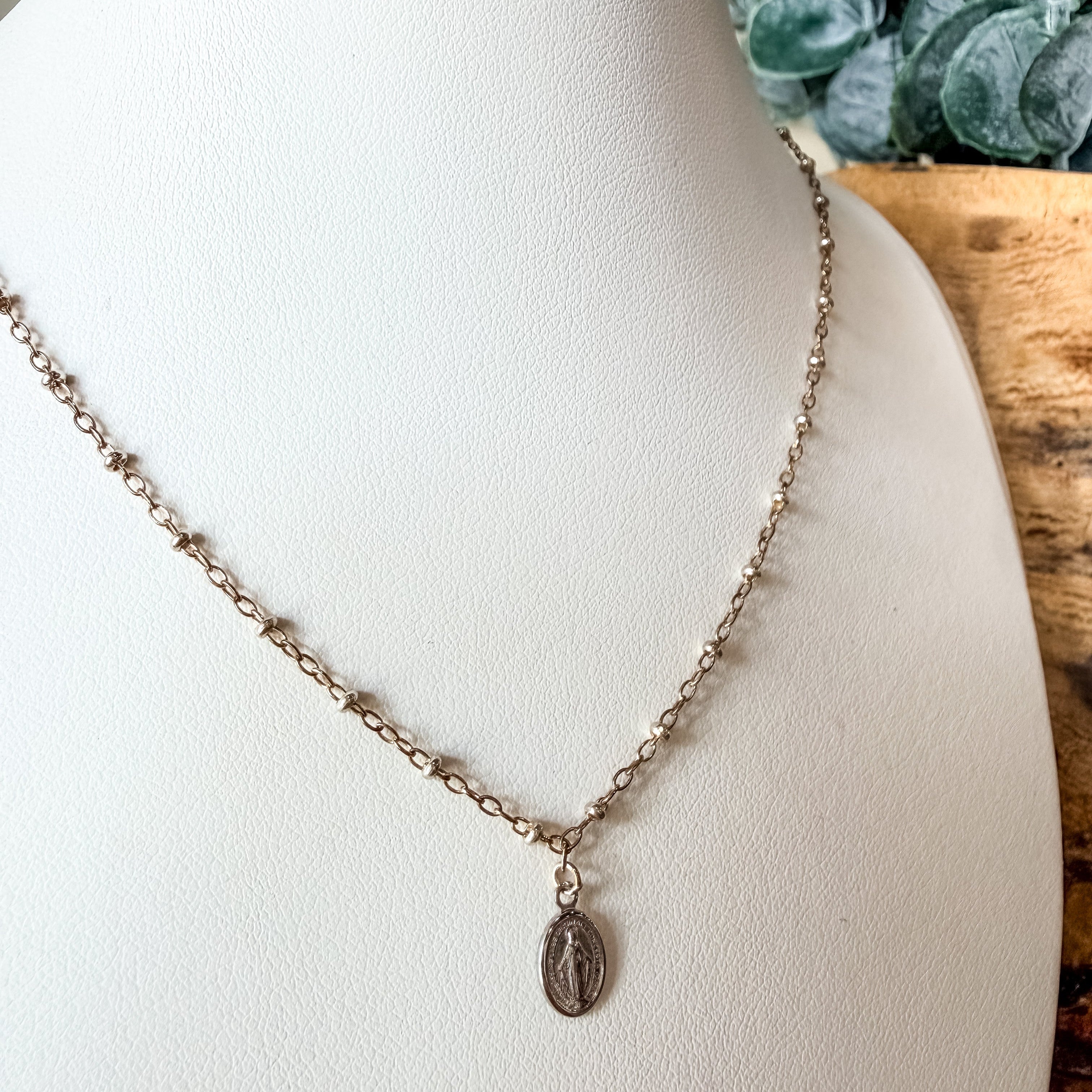 A delicate mixed metal chain necklace with a small oval religious pendant, displayed on a white jewelry bust.