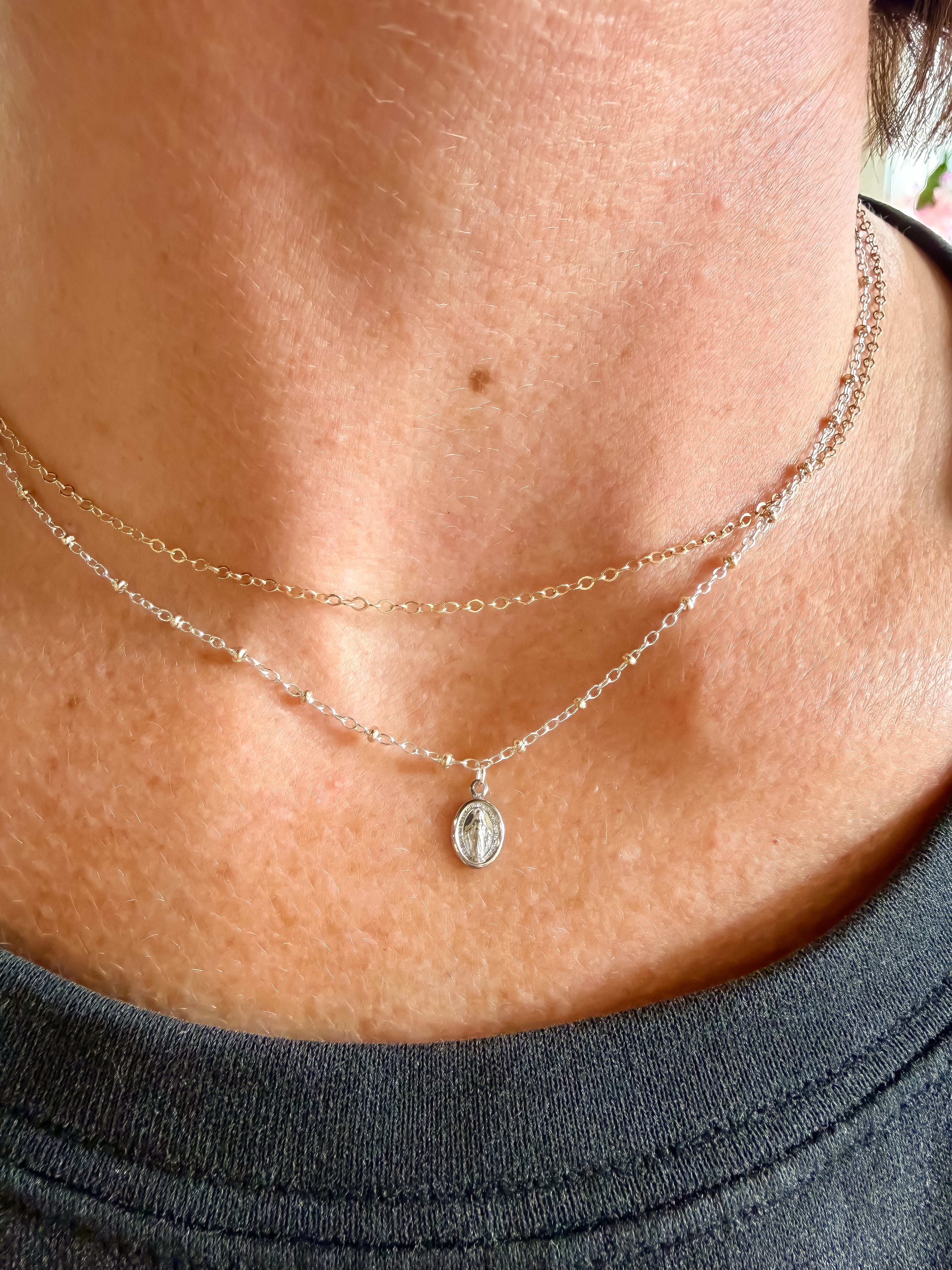 A close-up lifestyle shot of a woman wearing a delicate double-layered silver necklace with a small miraculous medal pendant on a sterling silver delicate cable chain. 