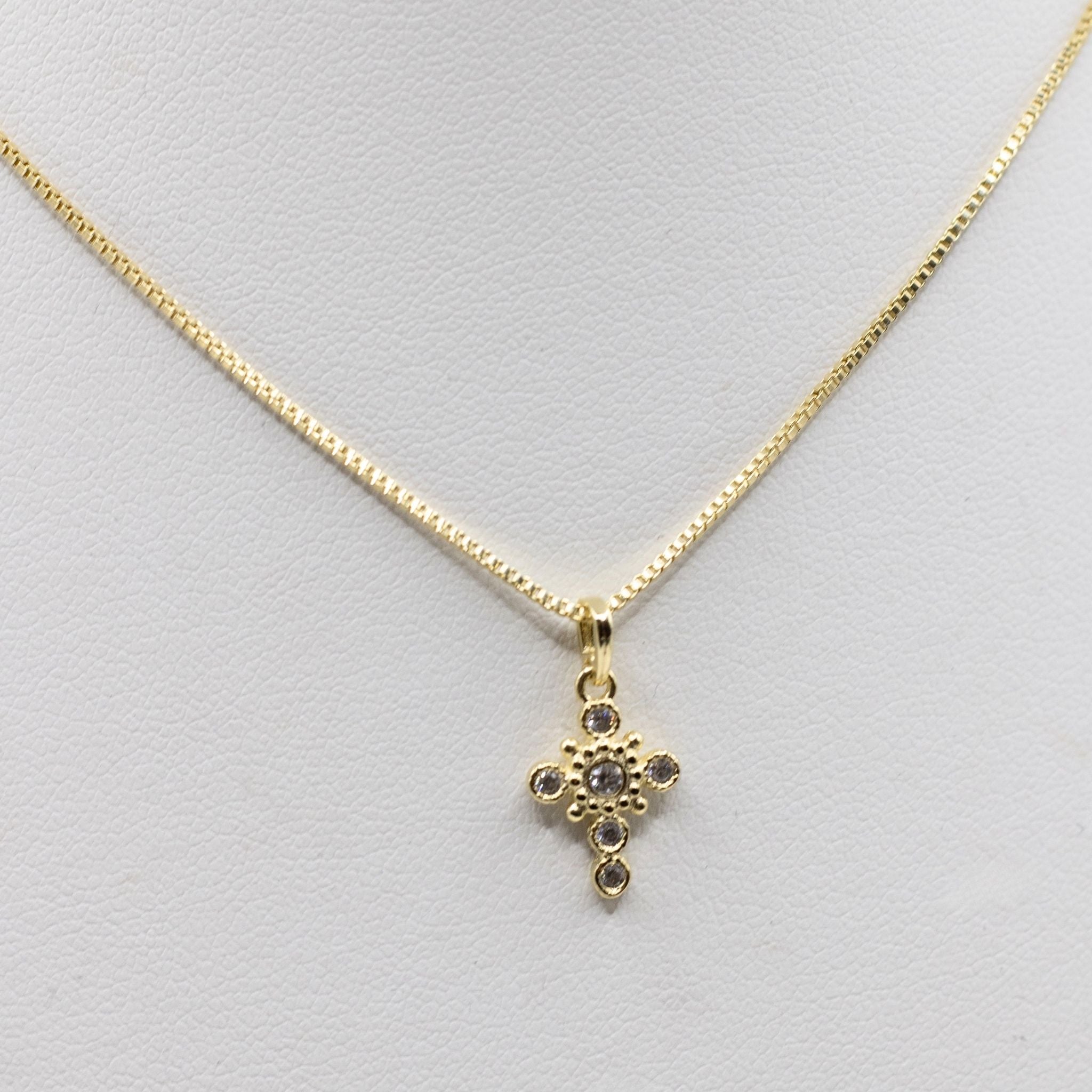 A gold cross pendant necklace with small clear stones, displayed on a white jewelry bust.